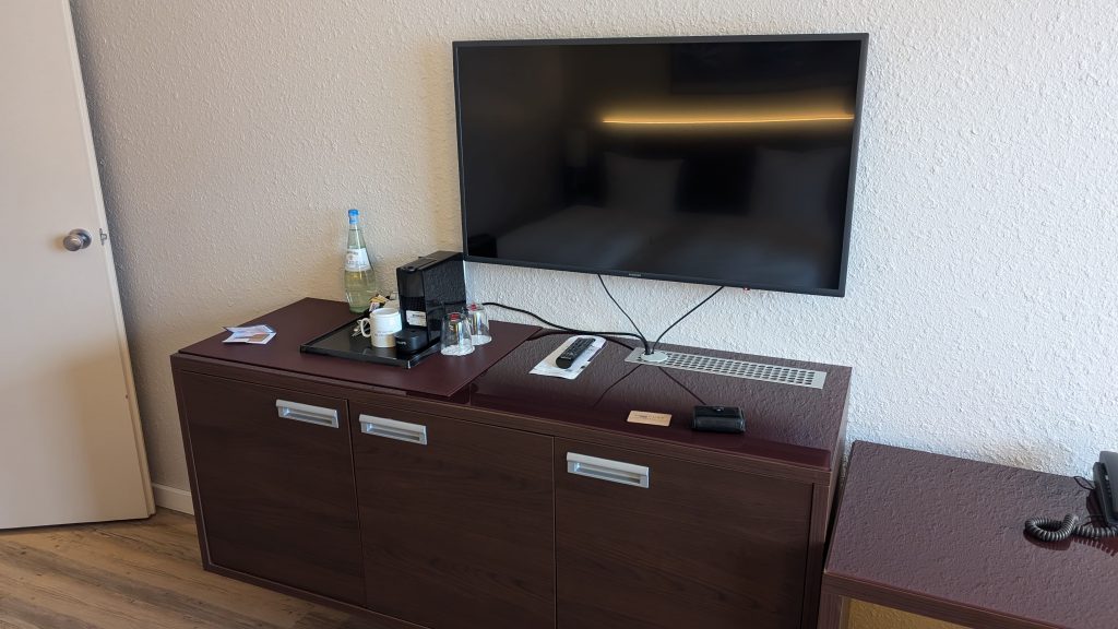 Die Bilder zeigen das Hotelzimmer und den Ausblick aus dem Fenster. Es gibt ein Doppelbett, einen Schreibtisch mit Schreibtischstuhl, einen Fernseher und eine Kommode.
