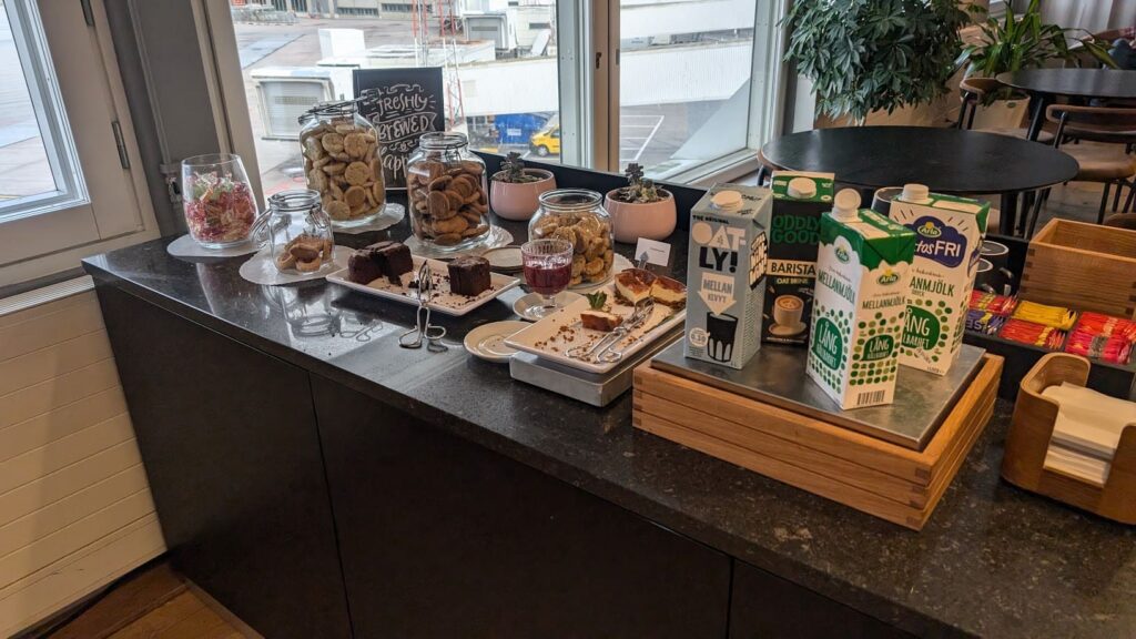 Dessertstation mit Keksgläsern, Kuchenstücken sowie Milch- und Haferdrinkpackungen vor dem Vorfeldfenster
